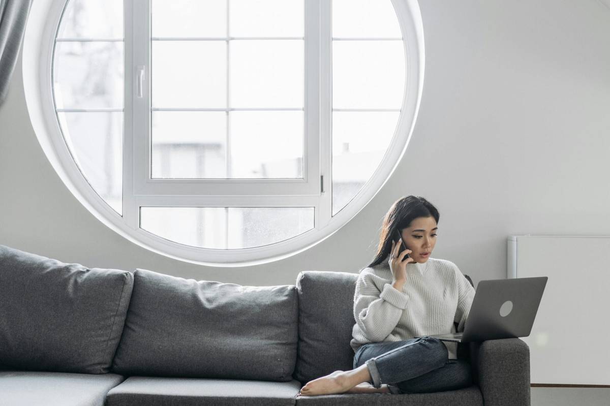 A woman sitting on a gray sofa, talking on the phone while using a laptop, with a large round window in the background.